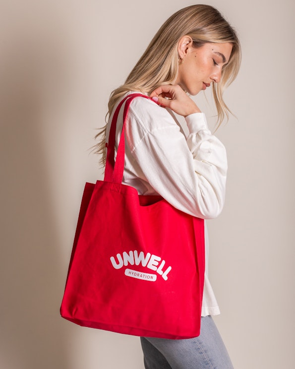 Screen printed red canvas tote bag with white arched brand logo and tagline, produced by MerchCamp for UNWELL