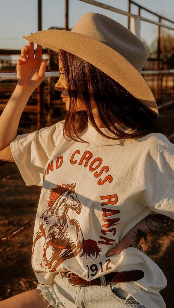 Woman in cowboy hat wearing vintage-style Sand Cross Ranch 1912 horse graphic tee outdoors, custom merchandise by MerchCamp