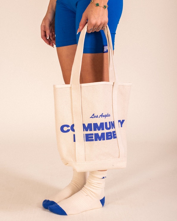 Screen printed cream canvas tote bag with bold blue "Community Member" text, produced by MerchCamp for Community Member