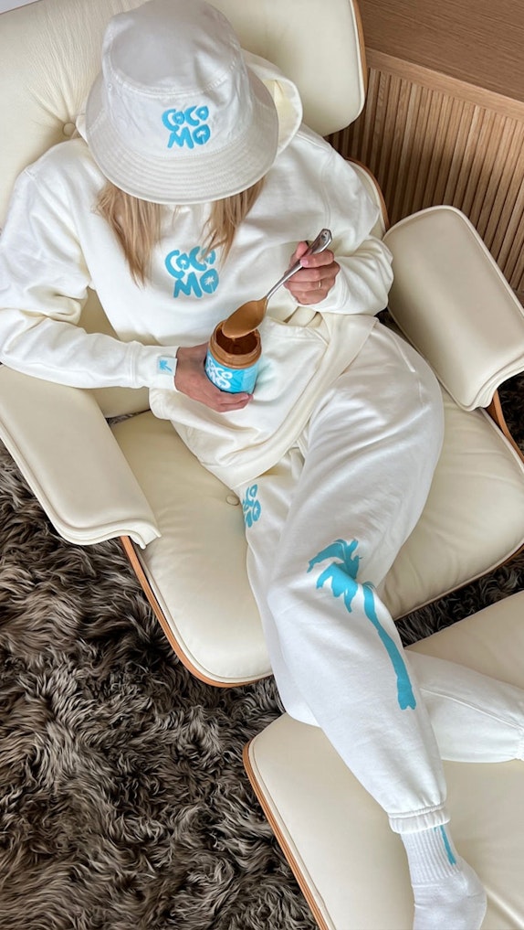 Person in white Coco Mo branded loungewear and bucket hat eating from a jar on leather chair — MerchCamp custom merchandise