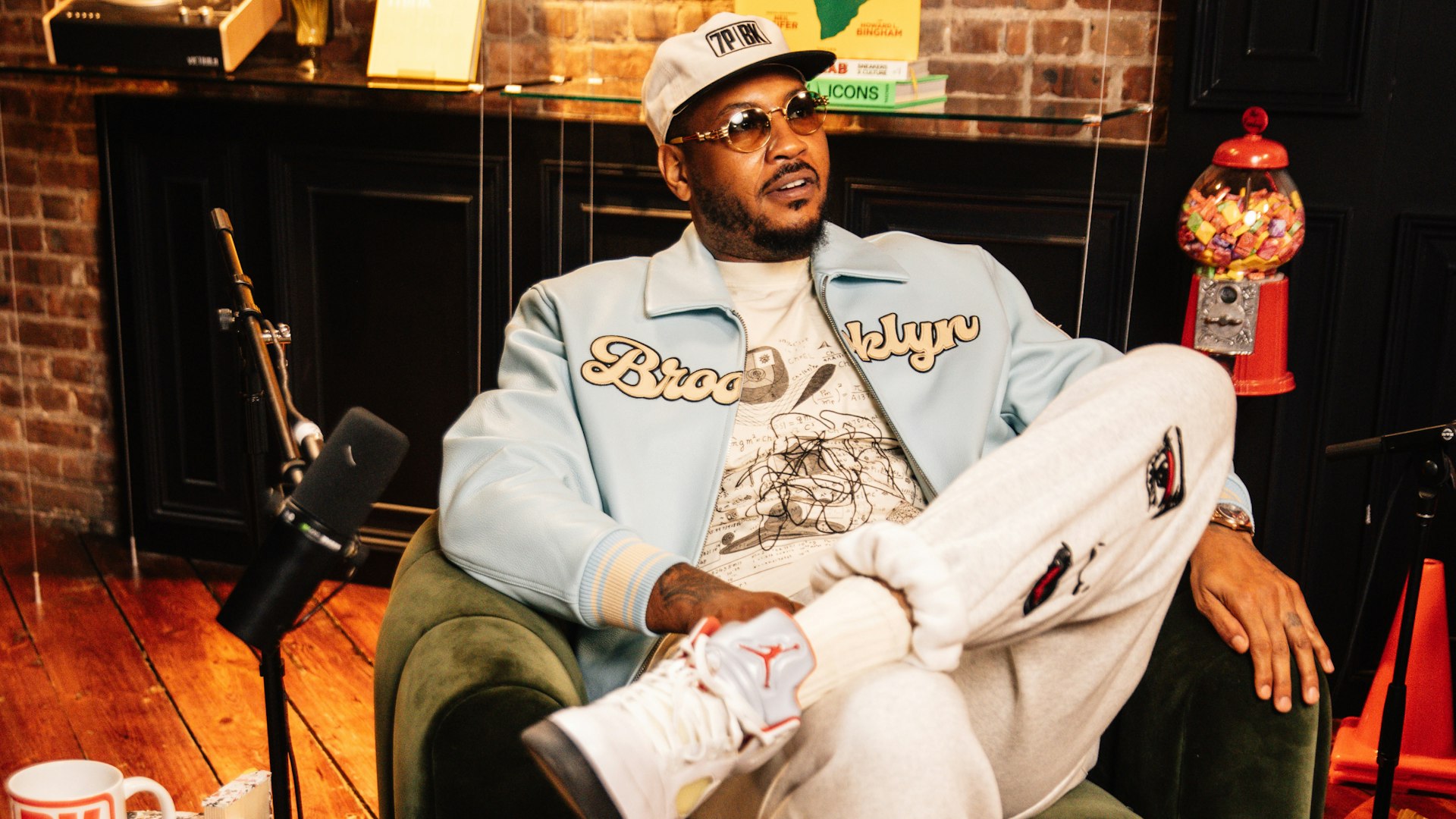 Man in Brooklyn varsity jacket and Jordan sneakers sitting in podcast studio — WAVE Sports + Entertainment merchandise by MerchCamp