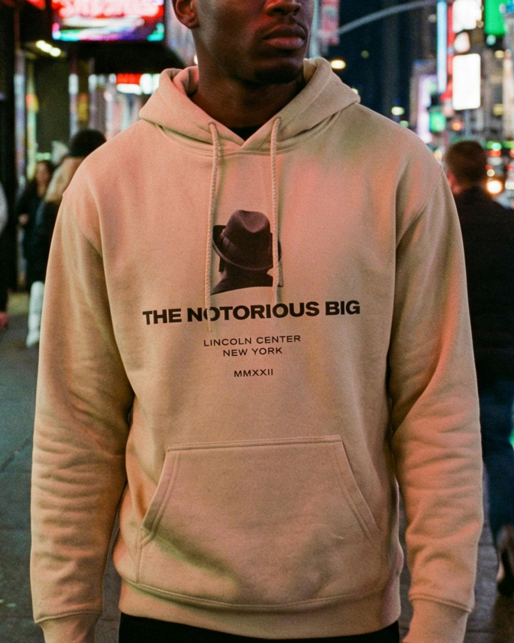 Man wearing beige Notorious BIG hoodie with fedora graphic on city street at night — Biggie Smalls merchandise by MerchCamp