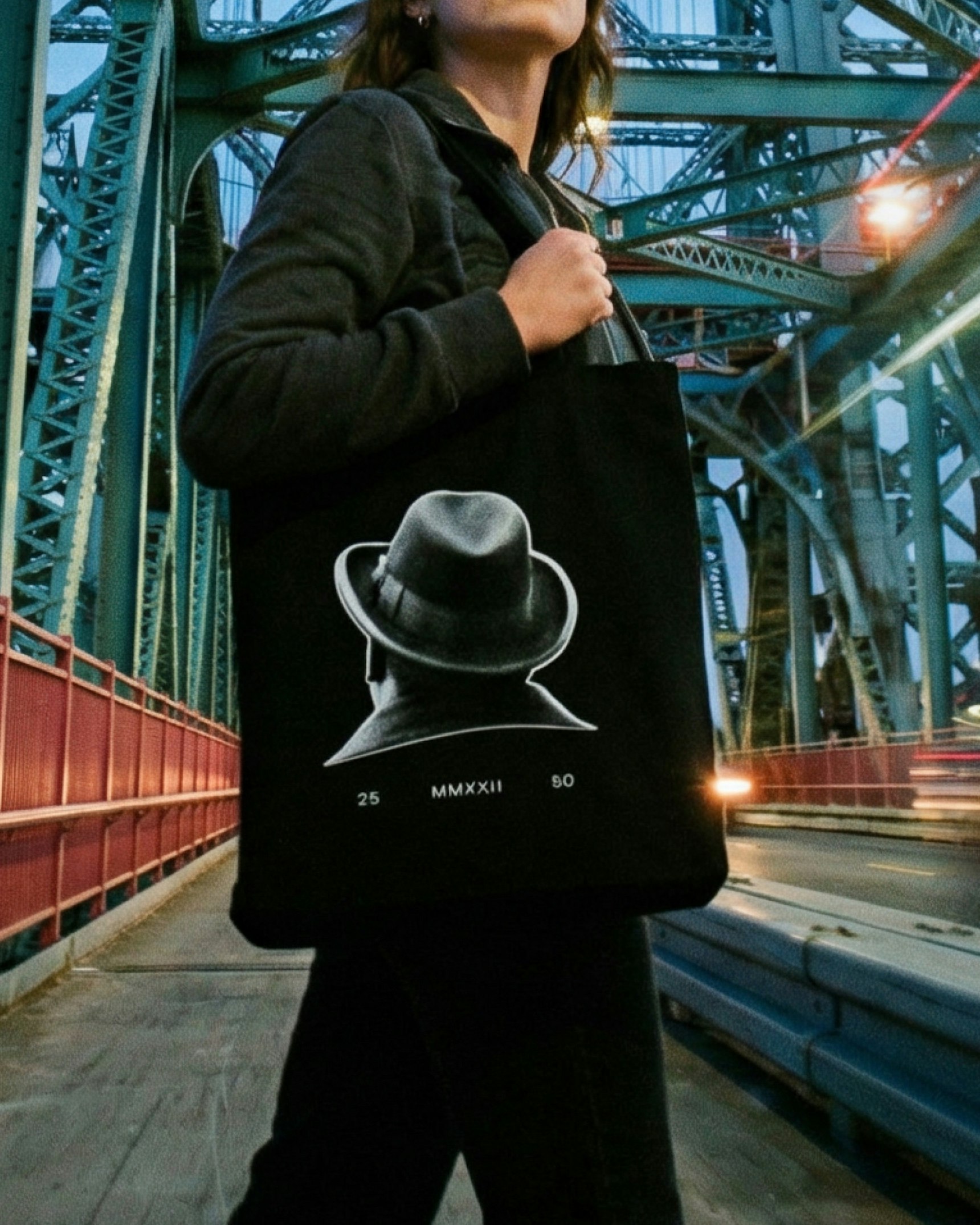 Woman holding black tote bag with fedora silhouette graphic on industrial bridge at dusk — Biggie Smalls merchandise by MerchCamp