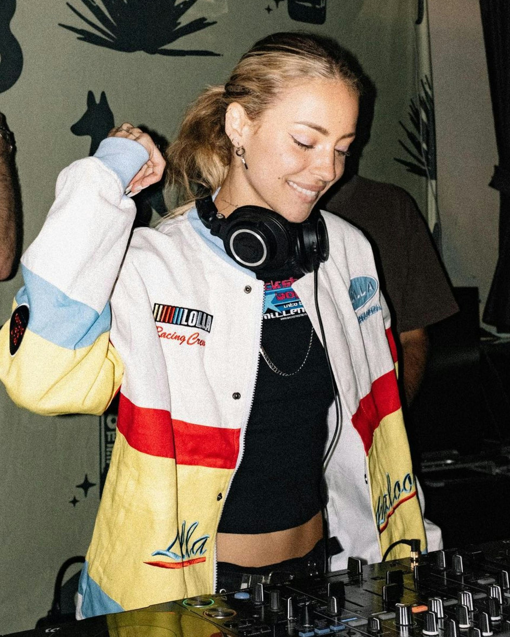 Smiling DJ wearing colorful Lolla Racing Crew jacket with headphones at mixer console — Lollapalooza merchandise by MerchCamp