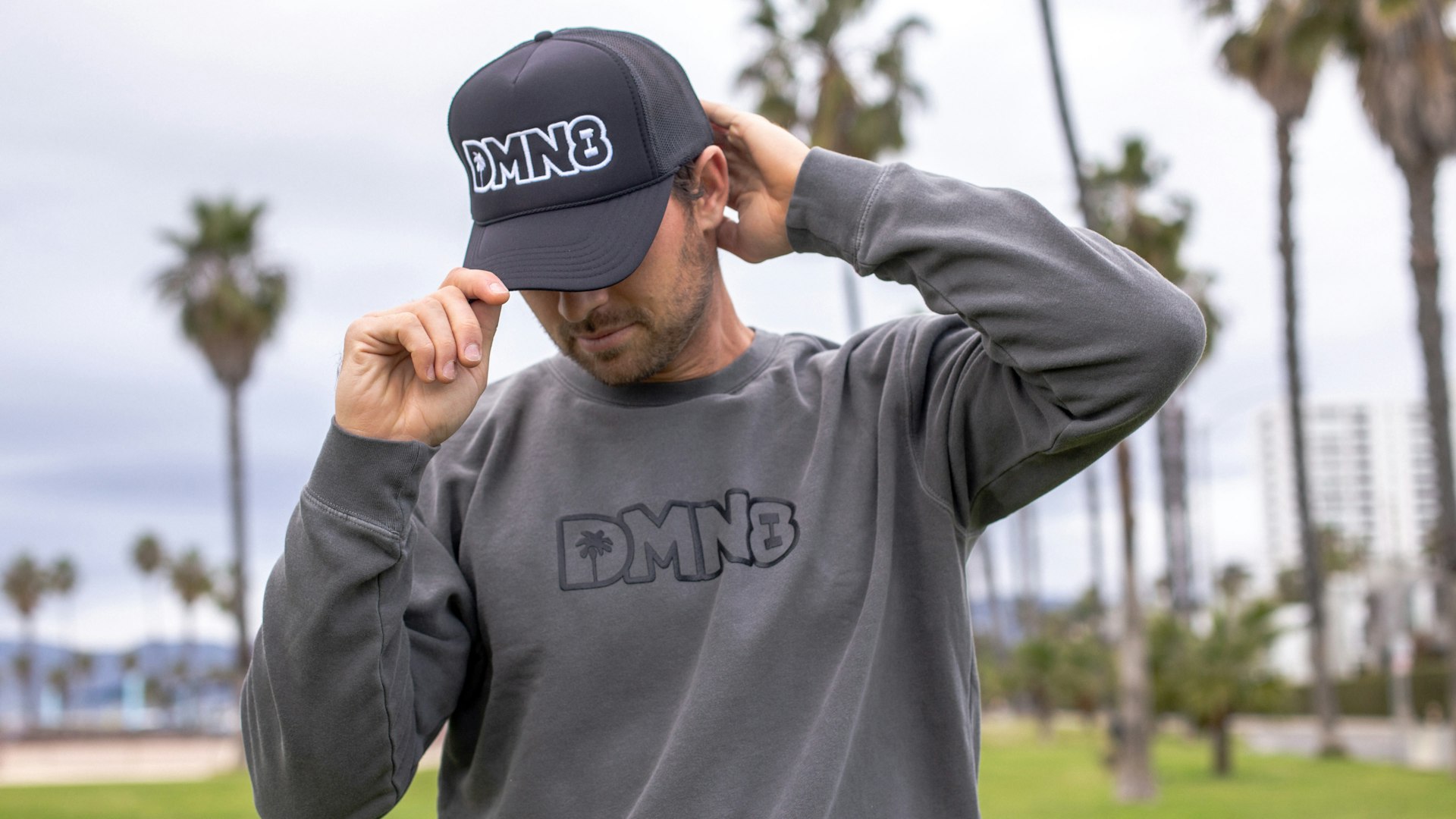 Man adjusting dark trucker cap and wearing matching grey DMN8 logo sweatshirt outdoors near palm trees — DMN8 merchandise by MerchCamp