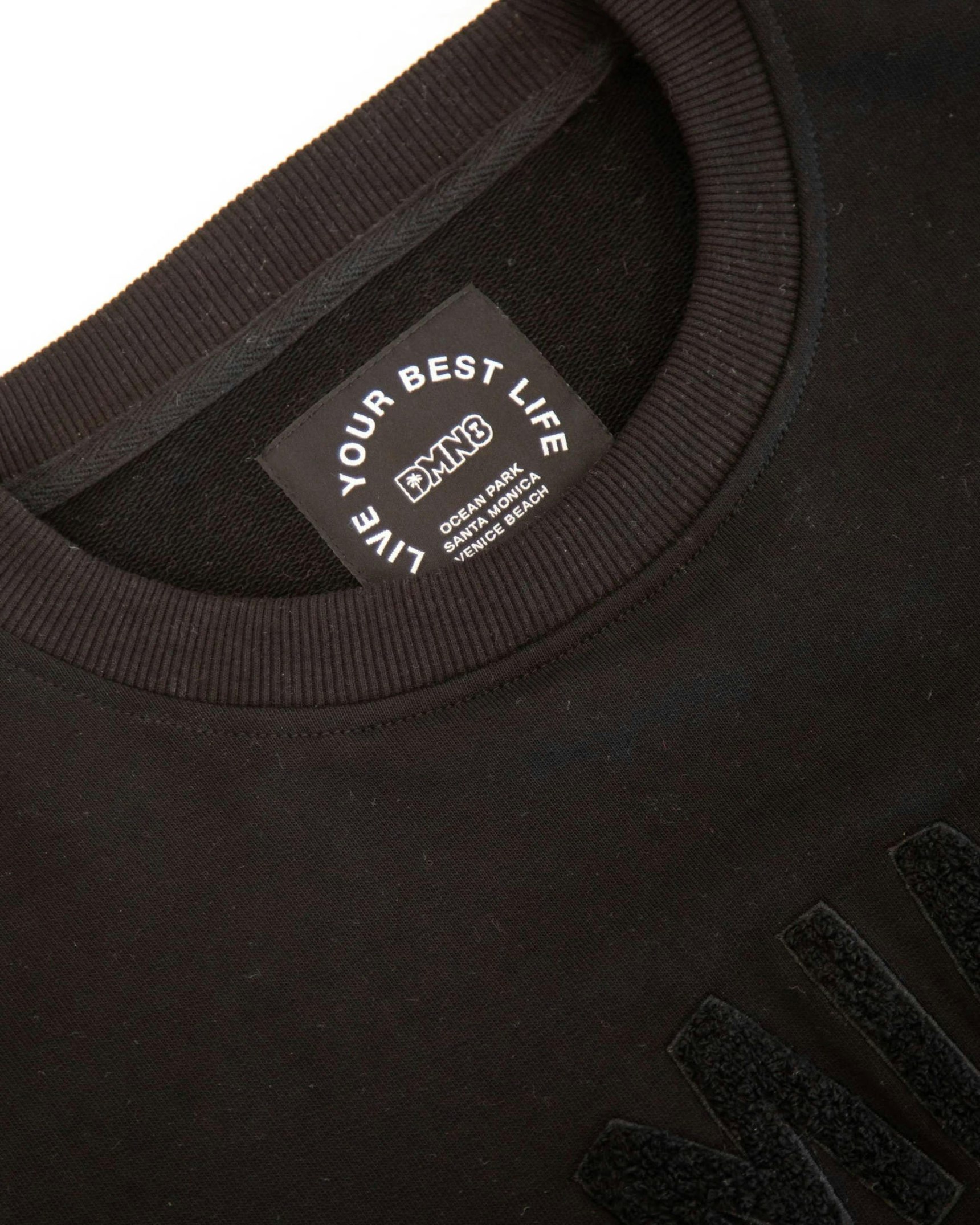 Close-up of black DMN8 crewneck sweatshirt showing woven neck label and chenille lettering detail — DMN8 merchandise by MerchCamp