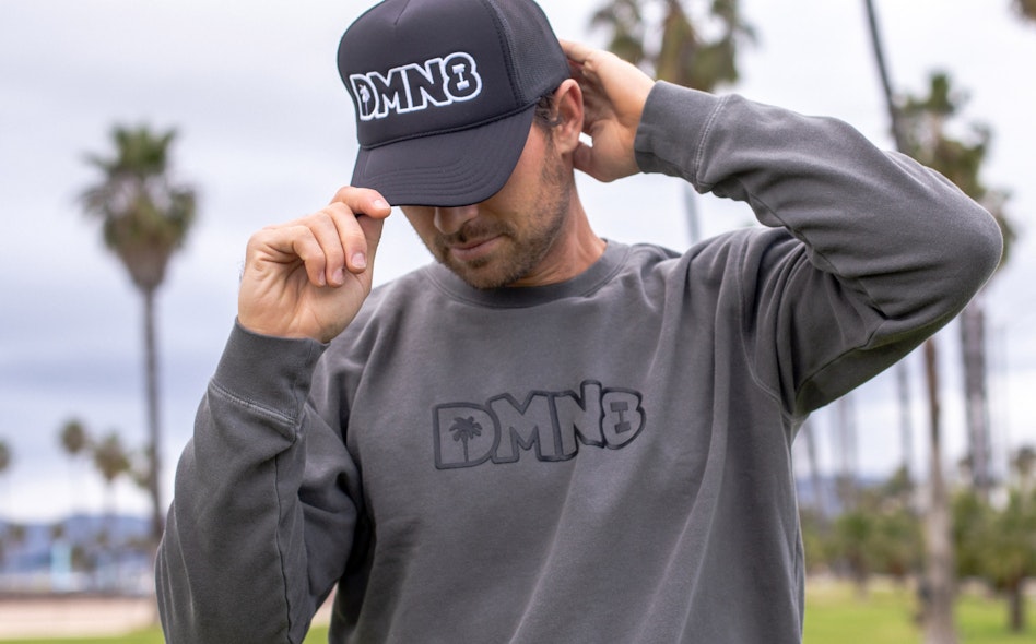 Man wearing DMN8 branded black trucker cap and charcoal crewneck sweatshirt with palm trees in background, produced by MerchCamp for DMN8