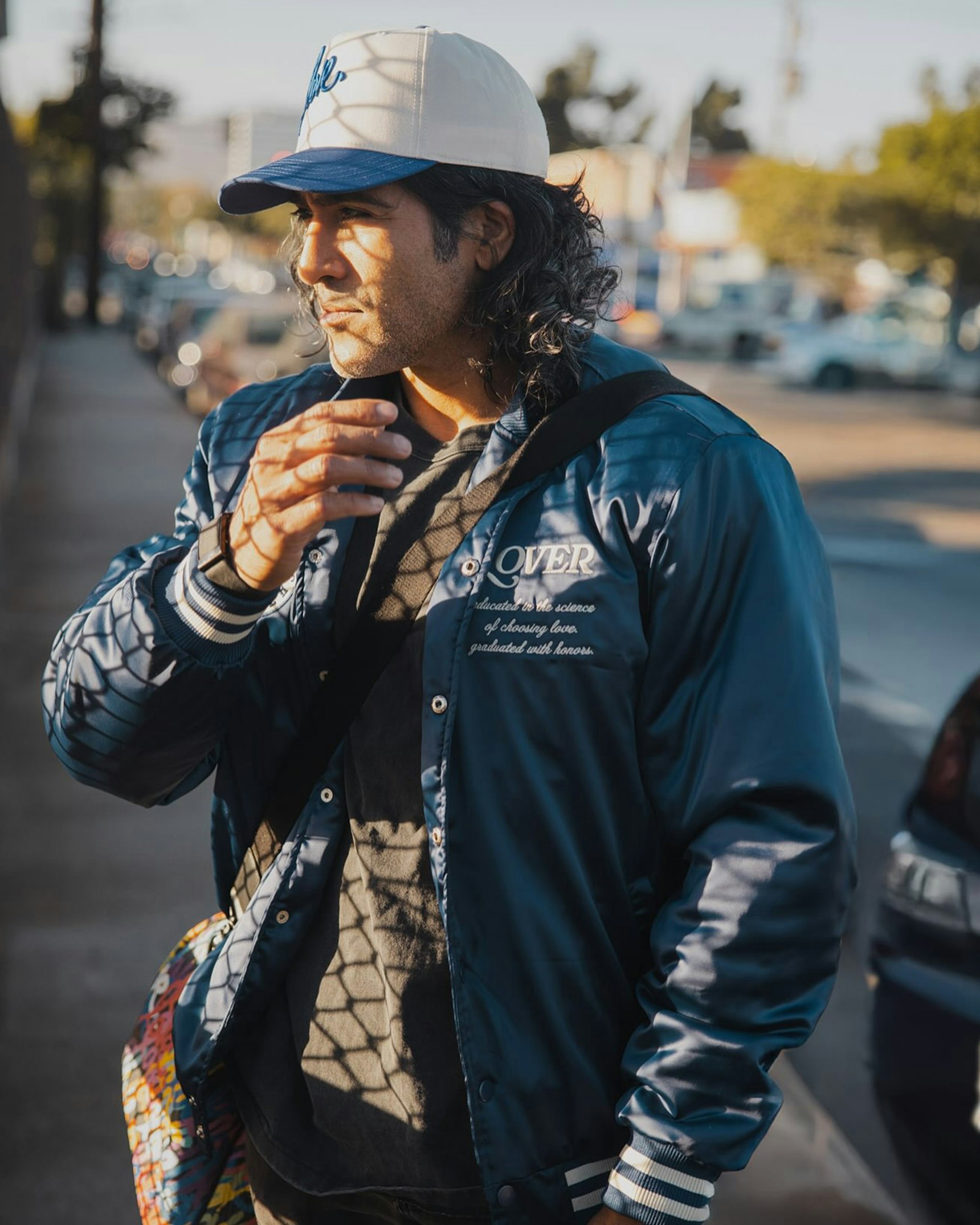 Man wearing blue satin "LOVER" bomber jacket and trucker cap on sunlit street — Ruben Rojas merchandise by MerchCamp