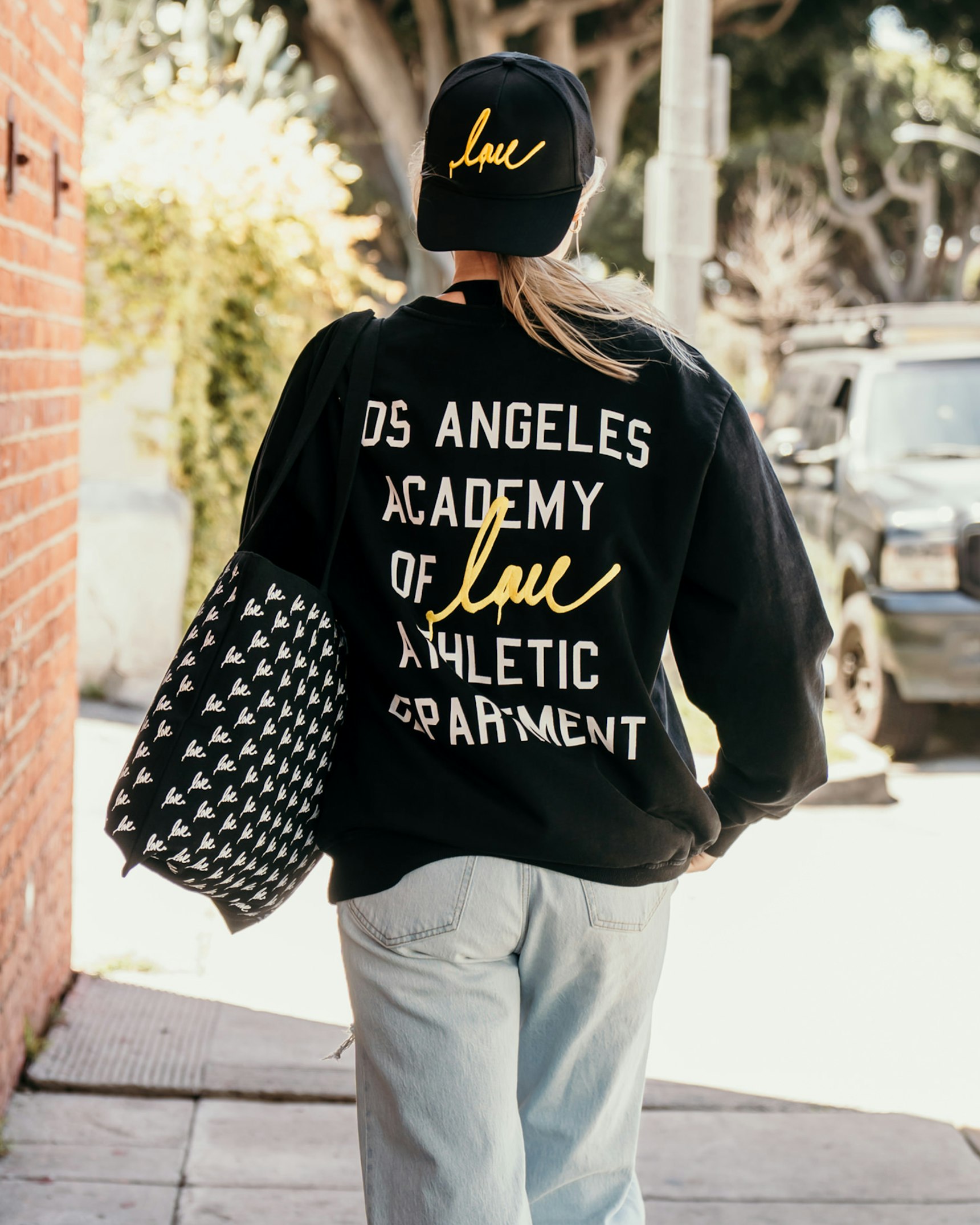Woman walking on sidewalk wearing black "Los Angeles Academy of Love Athletic Department" — Ruben Rojas merchandise by MerchCamp