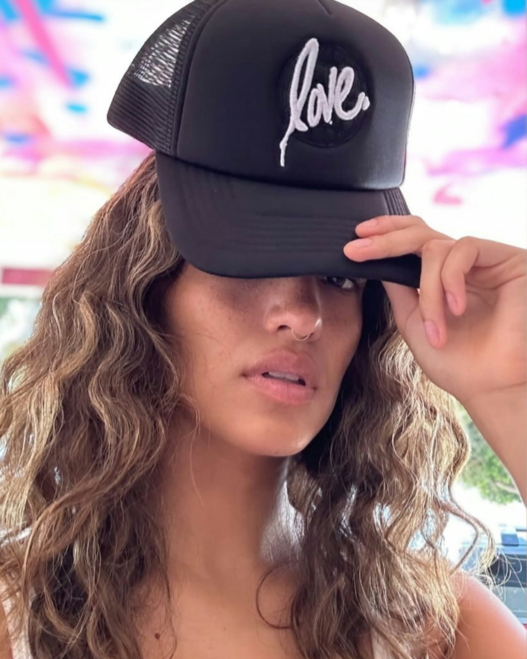 Woman with curly hair wearing black trucker hat with white "love." script, nose ring — Ruben Rojas merchandise by MerchCamp
