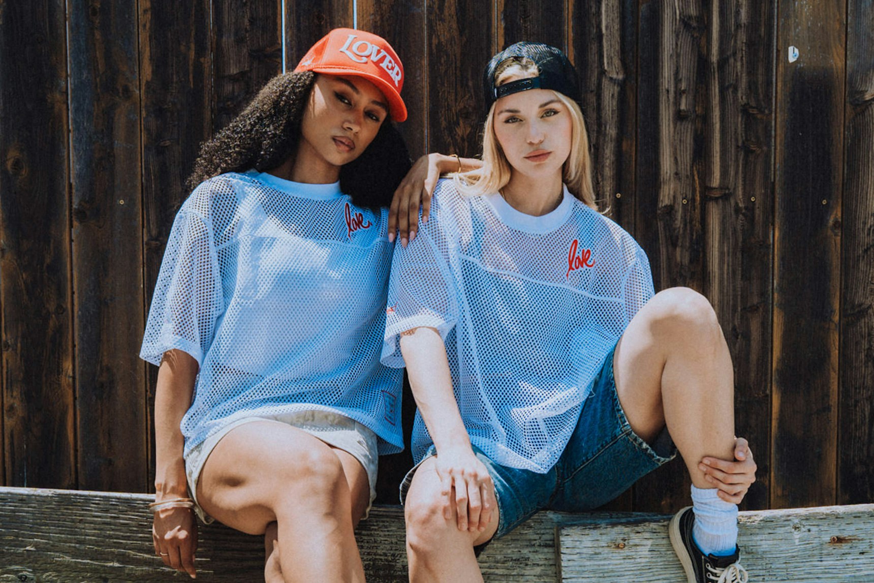 Two women in light blue mesh "Love" tees and caps sitting against dark wooden fence — Ruben Rojas merchandise by MerchCamp