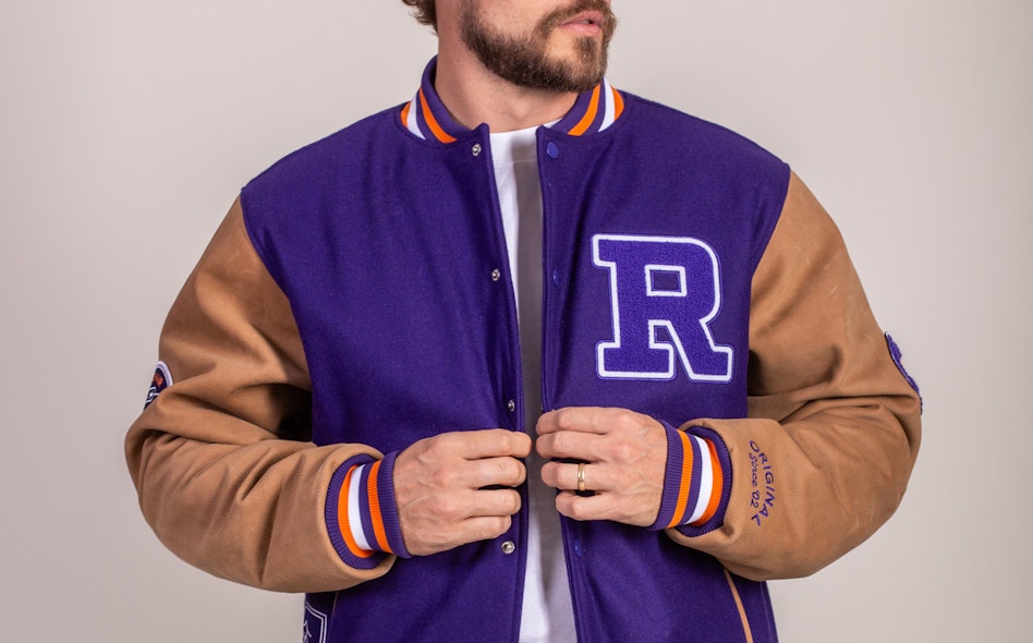 Purple wool varsity jacket with tan leather sleeves, chenille R patch, and orange-white striped ribbing, produced by MerchCamp for Roku
