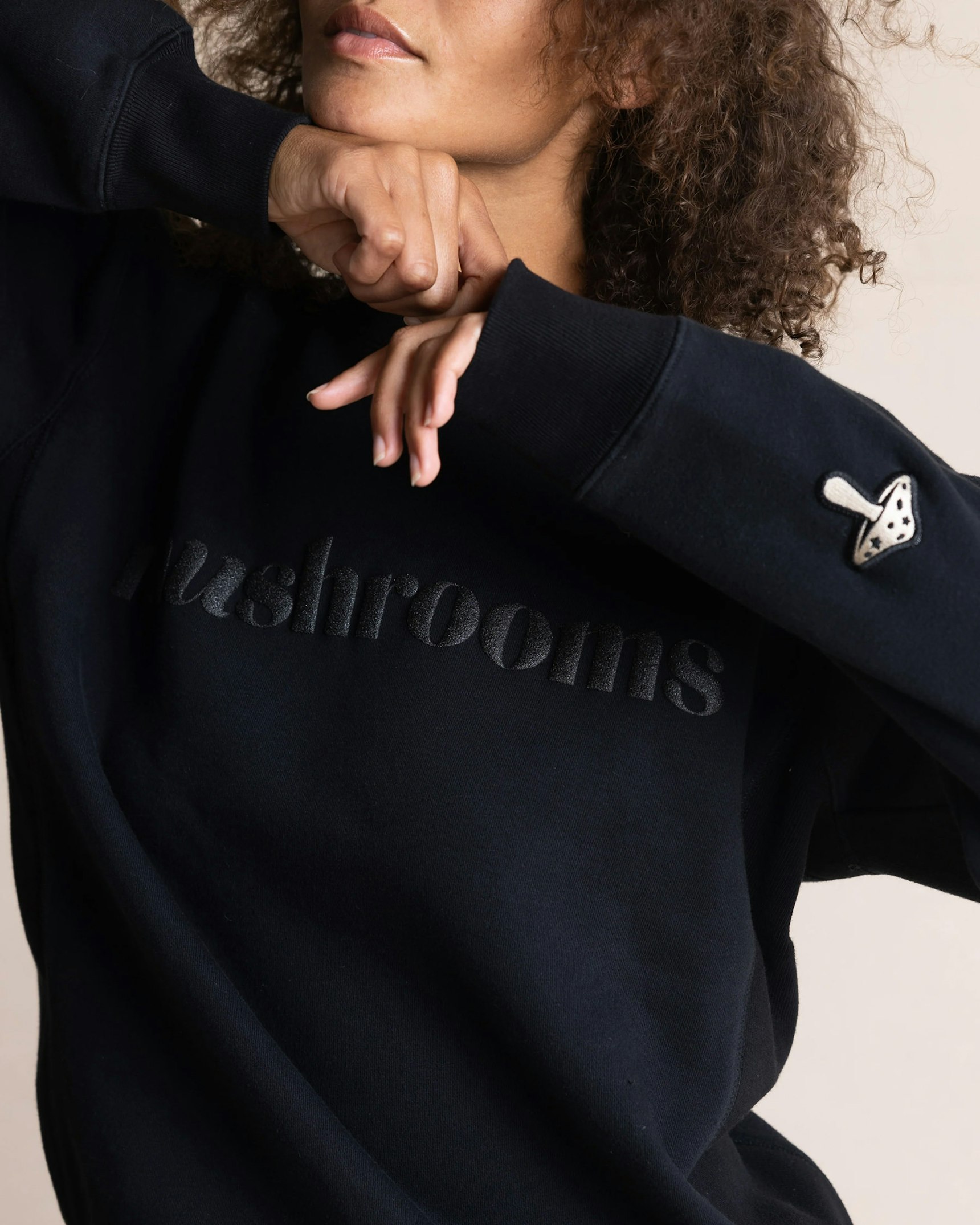 Woman in black crewneck sweatshirt with embossed "Mushrooms" text and mushroom sleeve patch — Alice Mushrooms merchandise by MerchCamp