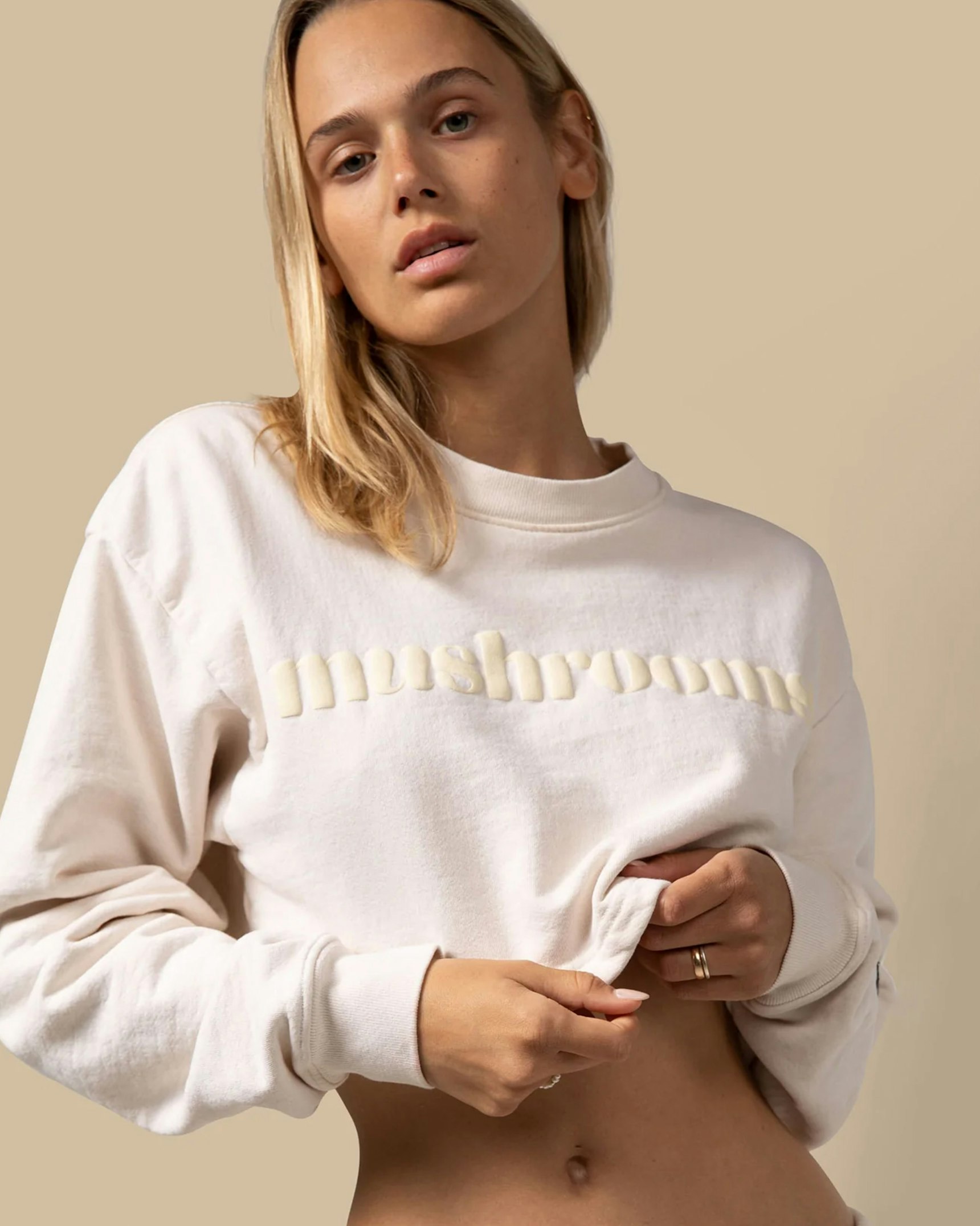 Woman in cream cropped sweatshirt with raised "mushrooms" lettering against beige backdrop — Alice Mushrooms merchandise by MerchCamp