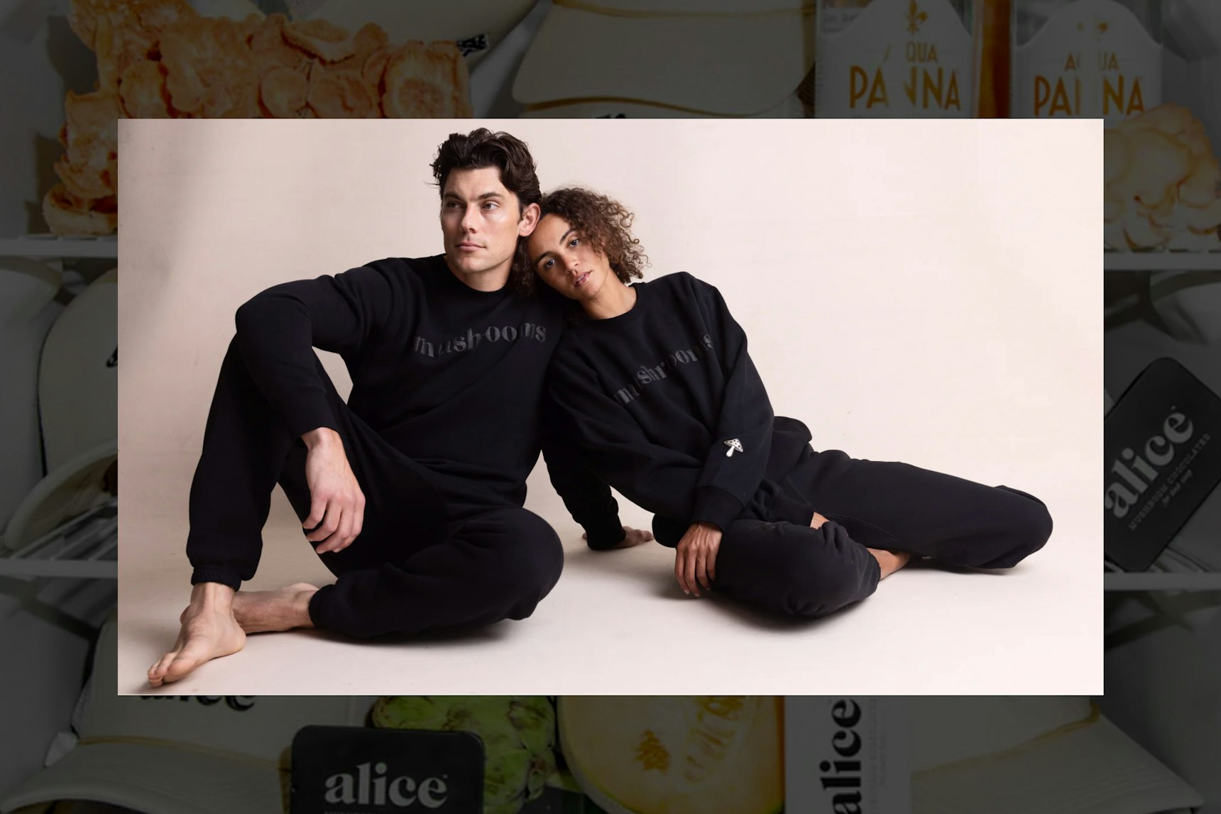 Two models in matching black "mushrooms" sweatsuit sets sitting on studio floor — Alice Mushrooms merchandise by MerchCamp