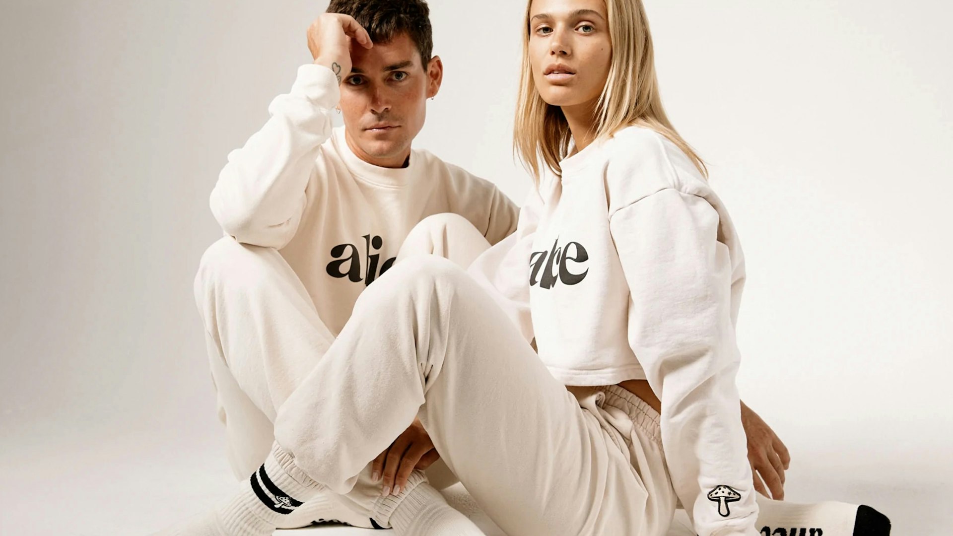 Male and female models sitting in matching cream Alice Mushrooms logo sweatsuit sets — Alice Mushrooms merchandise by MerchCamp