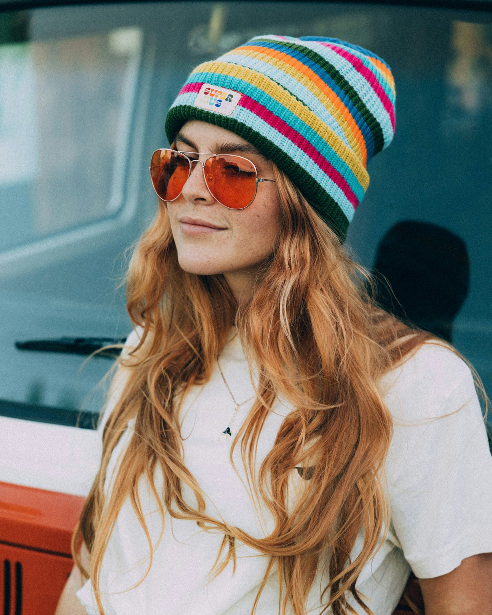 Woman with long red hair wearing colorful striped Supermush beanie, orange aviator sunglasses — Supermush merchandise by MerchCamp