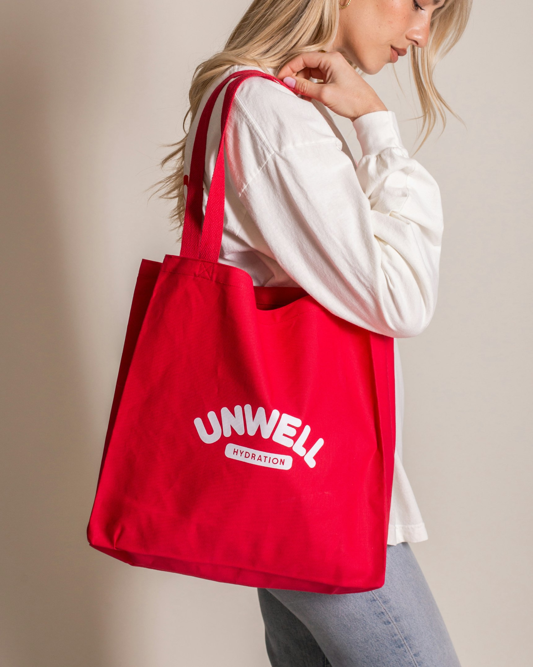 Blonde woman carrying red Unwell Hydration tote bag wearing white long-sleeve top and jeans — Unwell merchandise by MerchCamp