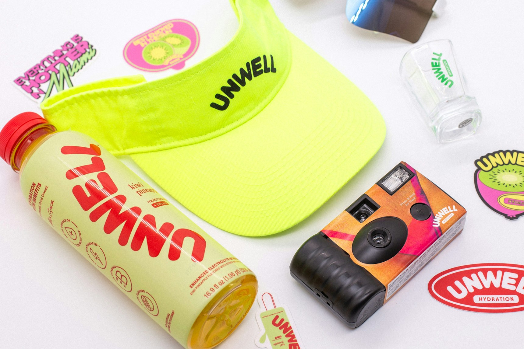 Flat lay of Unwell branded merchandise including neon visor, hydration drink, disposable camera — Unwell merchandise by MerchCamp