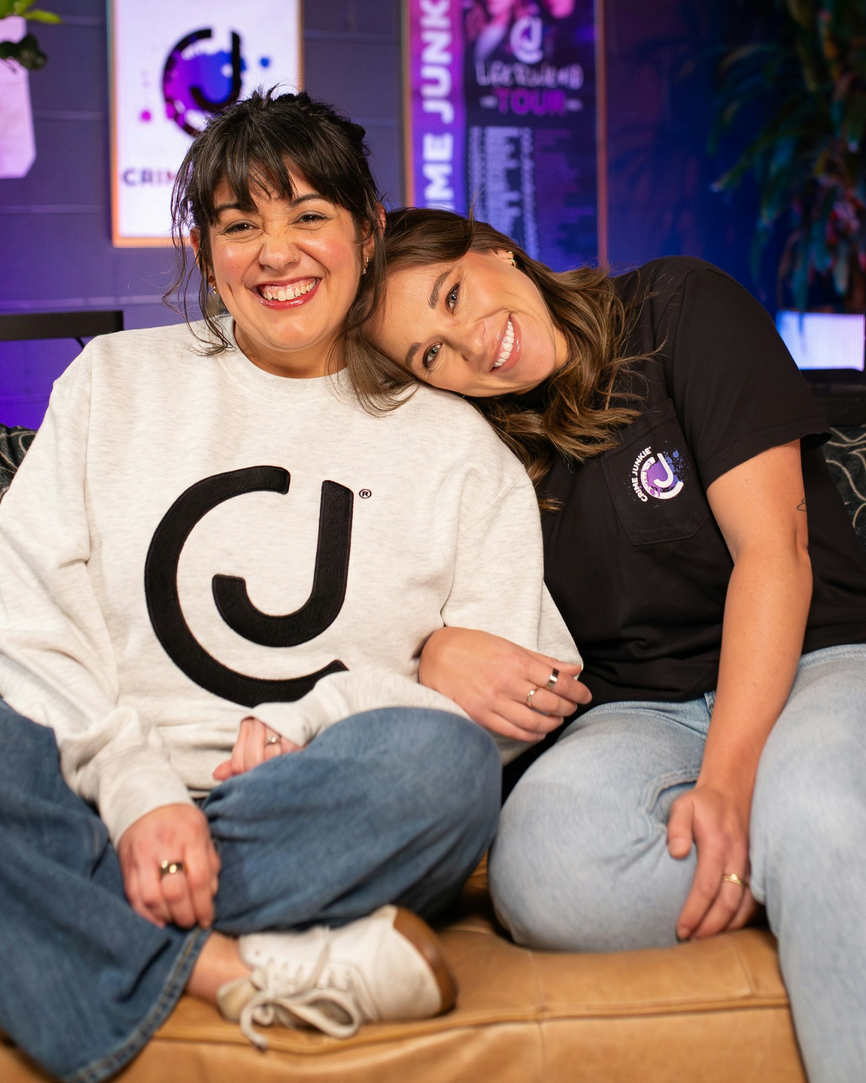 Two women in Crime Junkie branded merchandise sitting together on leather couch smiling — Crime Junkie merchandise by MerchCamp