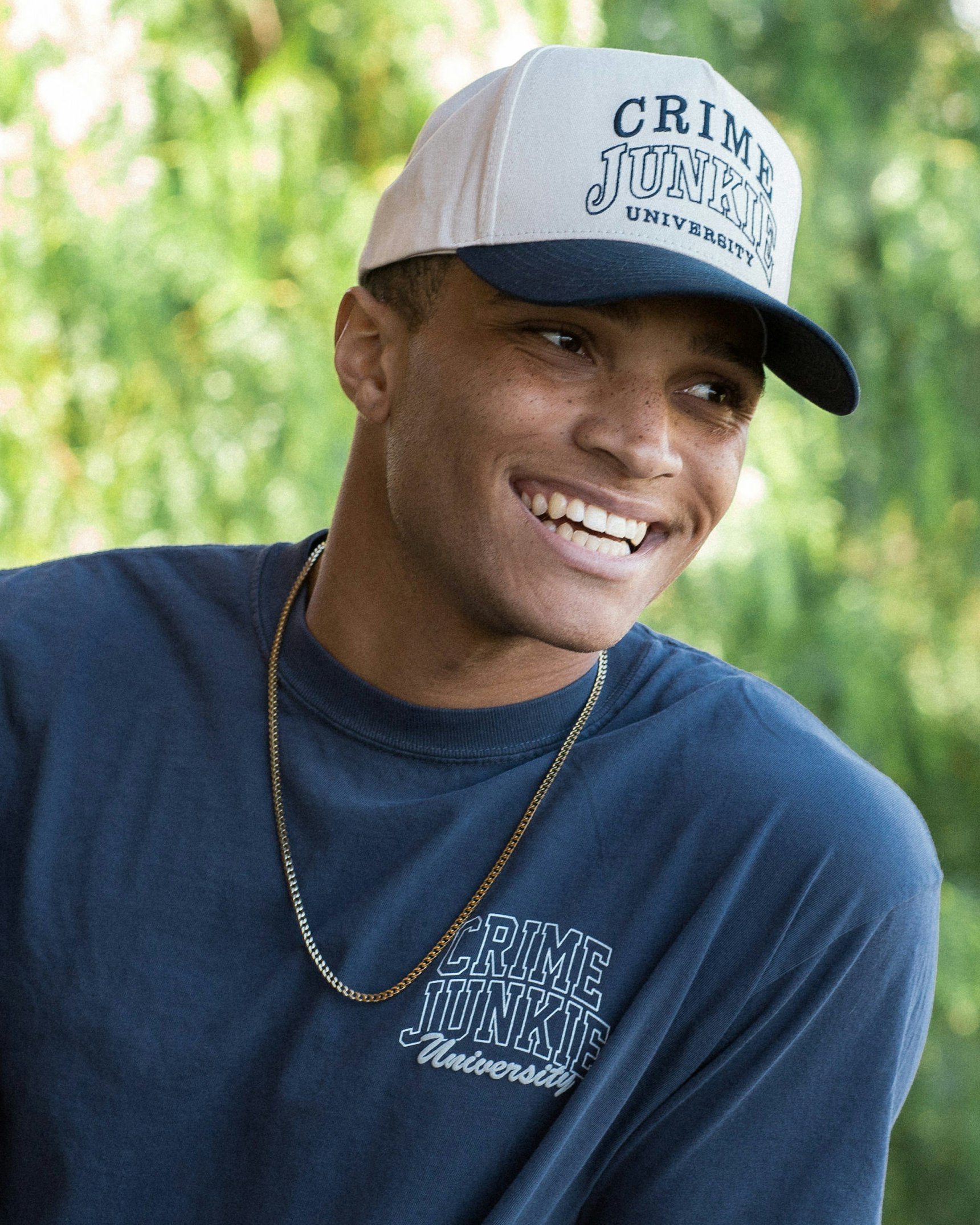 Smiling young man wearing navy Crime Junkie University trucker hat and matching t-shirt — Crime Junkie merchandise by MerchCamp