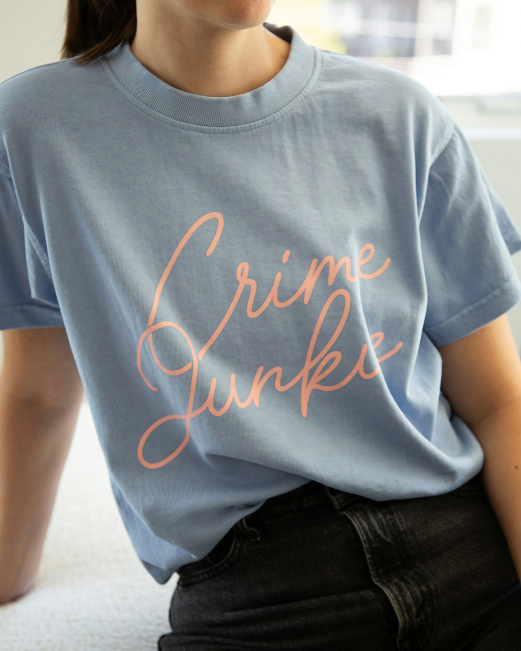 Woman wearing light blue Crime Junkie script t-shirt paired with dark denim jeans — Crime Junkie merchandise by MerchCamp
