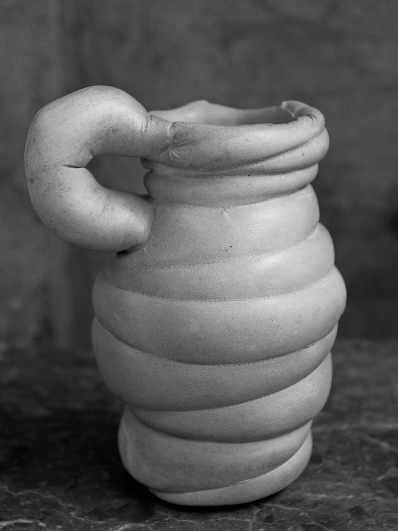 Black and white image displaying front view of Chub Vase I by Tessa Silva. It contains horizontal lines throughout the body and a handle. The material is made from the 'Chalk & Cheese' technique developed by manipulating proteins from fermented skimmed milk. This vase is exclusively made for the new 2024 spring collection 'Observations' by online gallery Metamorphoses Objects. 