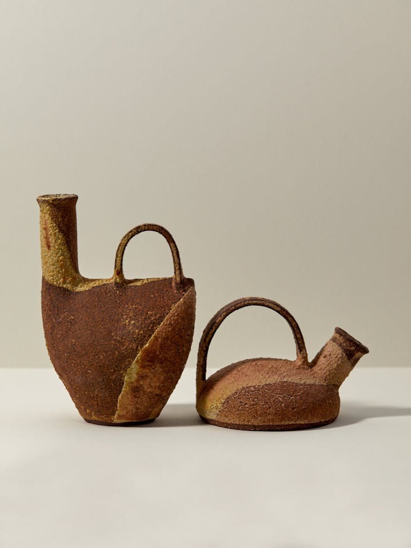 a set of two stoneware ceramic vessels by Catherine Dix with caramel, brown, cream and rust red colours, each vessel has an earthy texture includes a handle and pouring spout.
