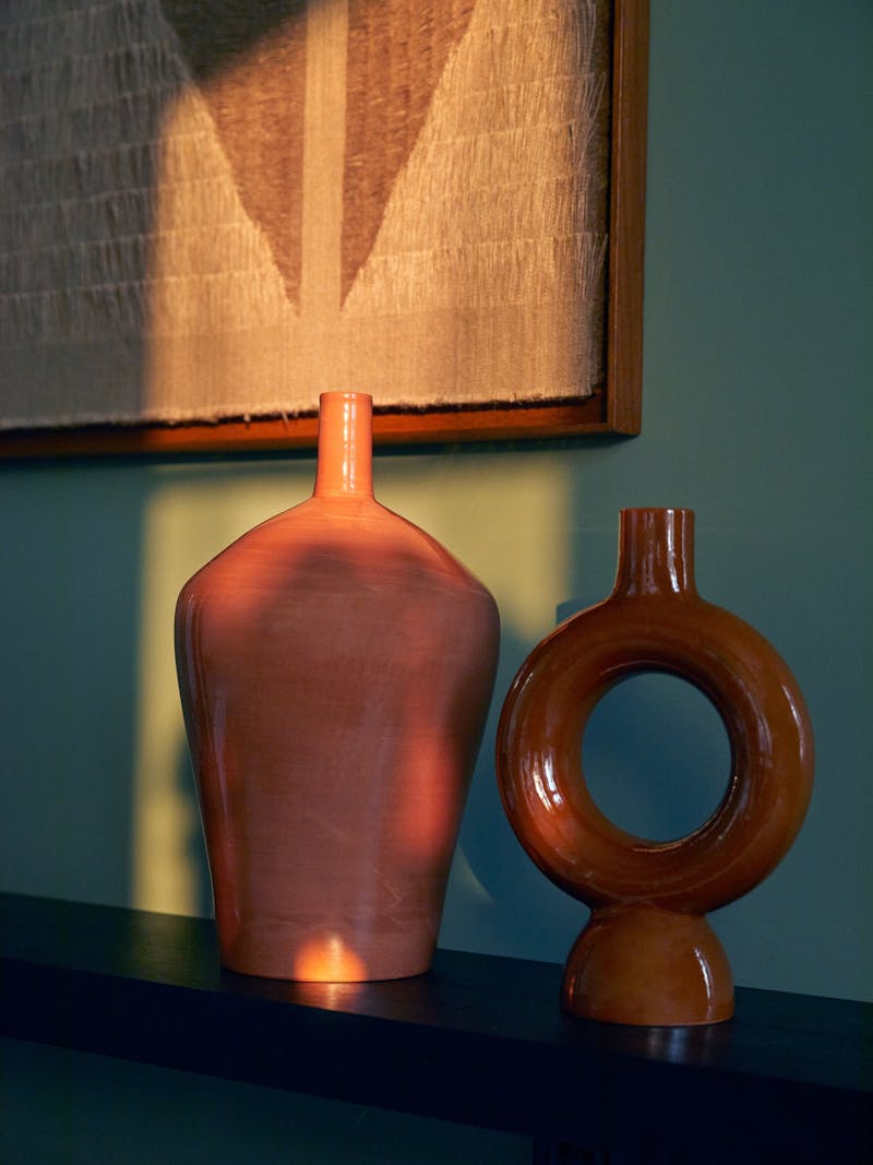 a pair of terracotta vessels next to each other on a wooden cabinet with a work by Sophie Rowley in the background. Both vessels are terracotta with a hand applied clear glaze.