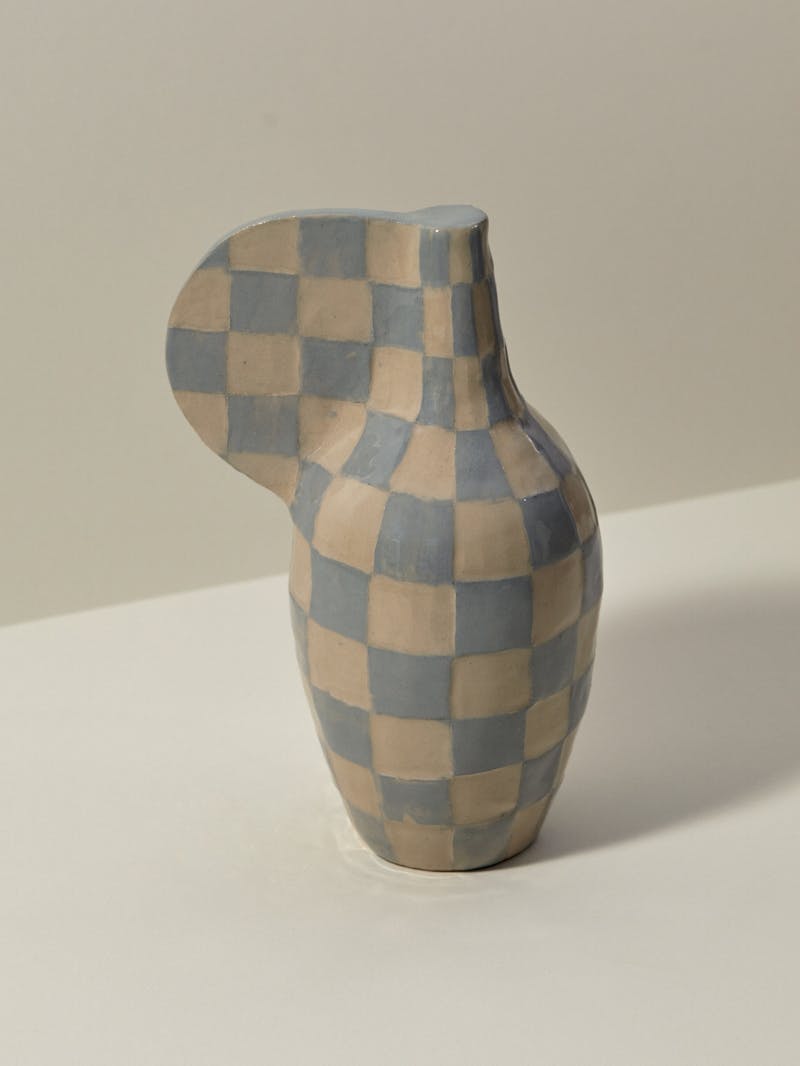 checkered and glazed stoneware vessel in blue and beige. The object is in the shape of a vase and has a solid tab like handle made by artist and ceramicist Maria Lenskjold.