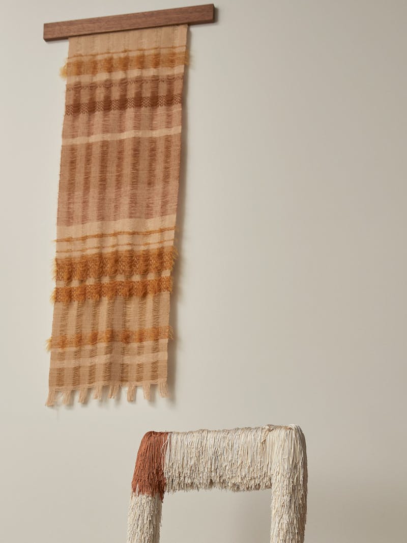 A hand woven wall hanging textile by Wovenform with colours of orange, pink, brown and cream. Part of a chair by Buro Belen is shown in the image. 