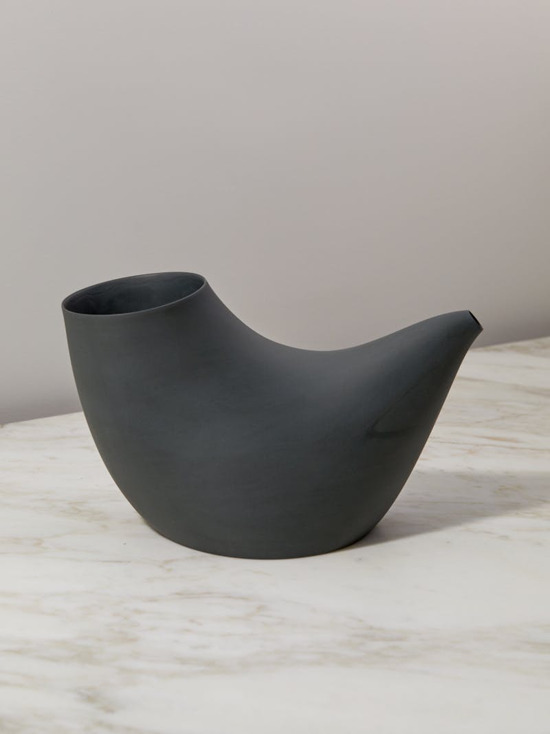 dark grey charcoal coloured porcelain vessel by Aldo Bakker. The pipe shaped container sits on a marble surface with a white background. 