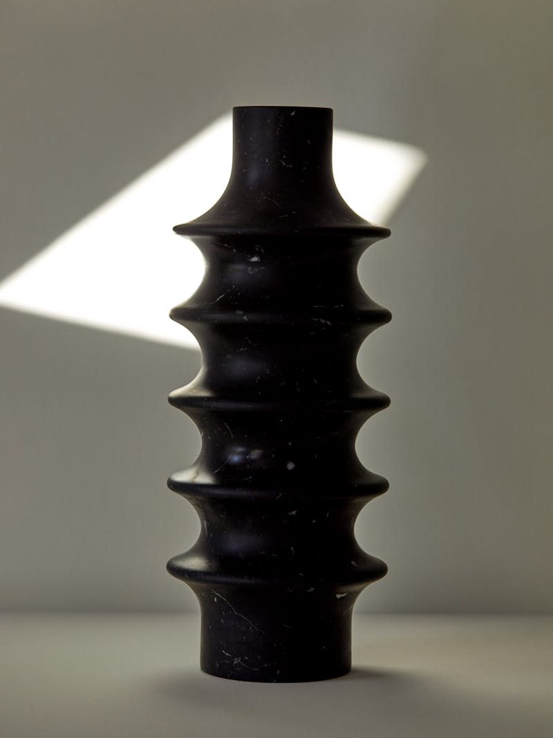 A hand carved stone vessel in black marquina marble sits on a white surface. The vase was crafted by Bloc Studios and has a unique ribbed cylindrical design. 