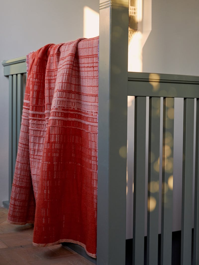 a pink, red and orange gradient mohair plaid blanket with linear and graphic stitching and weaving lays over a light green coloured stair rail. 