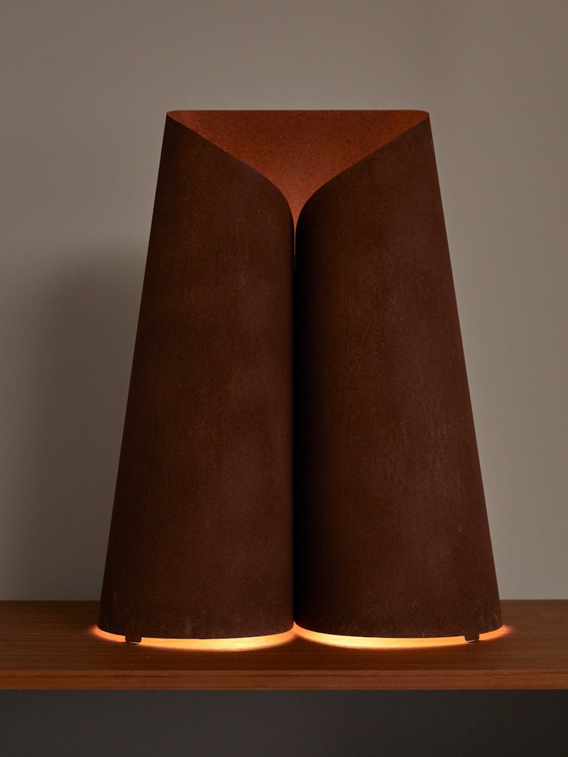 a lit up image of a hand-made bent rusted steel sheet metal lamp by Maria Tyakina. The lamp sits on a wooden table top and the light comes from the bottom.