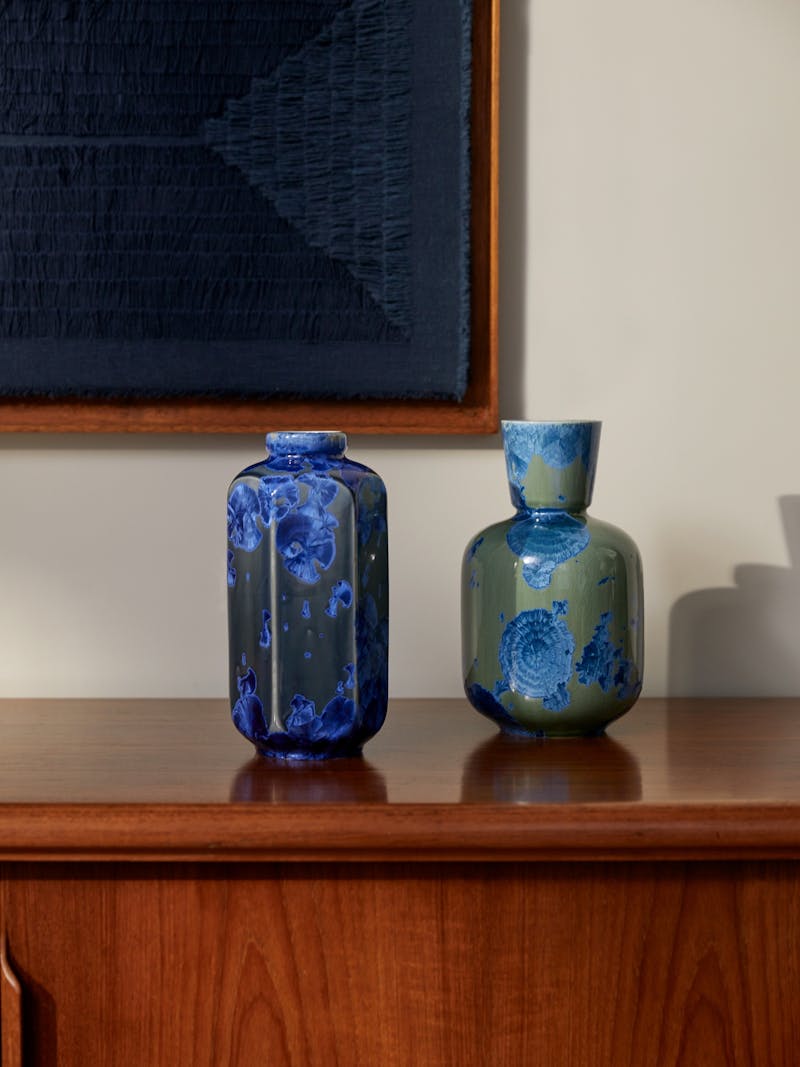 Two blue and green vases with a crystalline glaze sit on a cherry coloured wood cabinet. Each vase is hand made and glaze by artist Milan Pekar. 