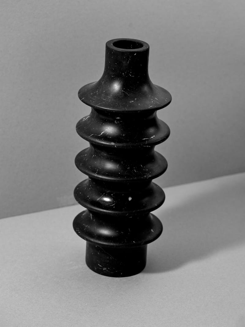 A hand carved stone vessel in black marquina marble sits on a white surface. The vase was crafted by Bloc Studios and has a unique ribbed cylindrical design. 