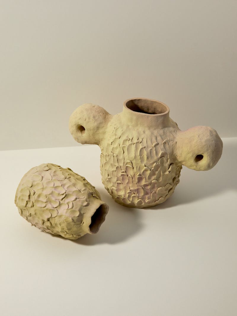 Two pinch pot style stoneware ceramic glazed vessels from French artist Pauline Bonnet, one sits upright and has two handles and the other is laid down on the surface of the table. 