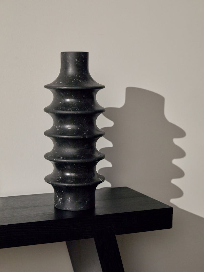 A hand carved stone vessel in black marquina marble sits on a black bench. The vase was crafted by Bloc Studios and has a unique ribbed cylindrical design. 