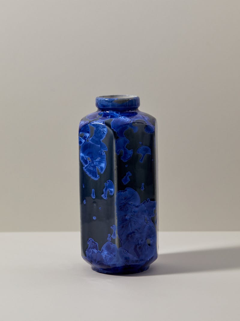 A hand made hexagonal porcelain vase sits on a table top, the body of the vase is in a dark blue and there is a royal blue crystalline geode like glaze over the top of it. 