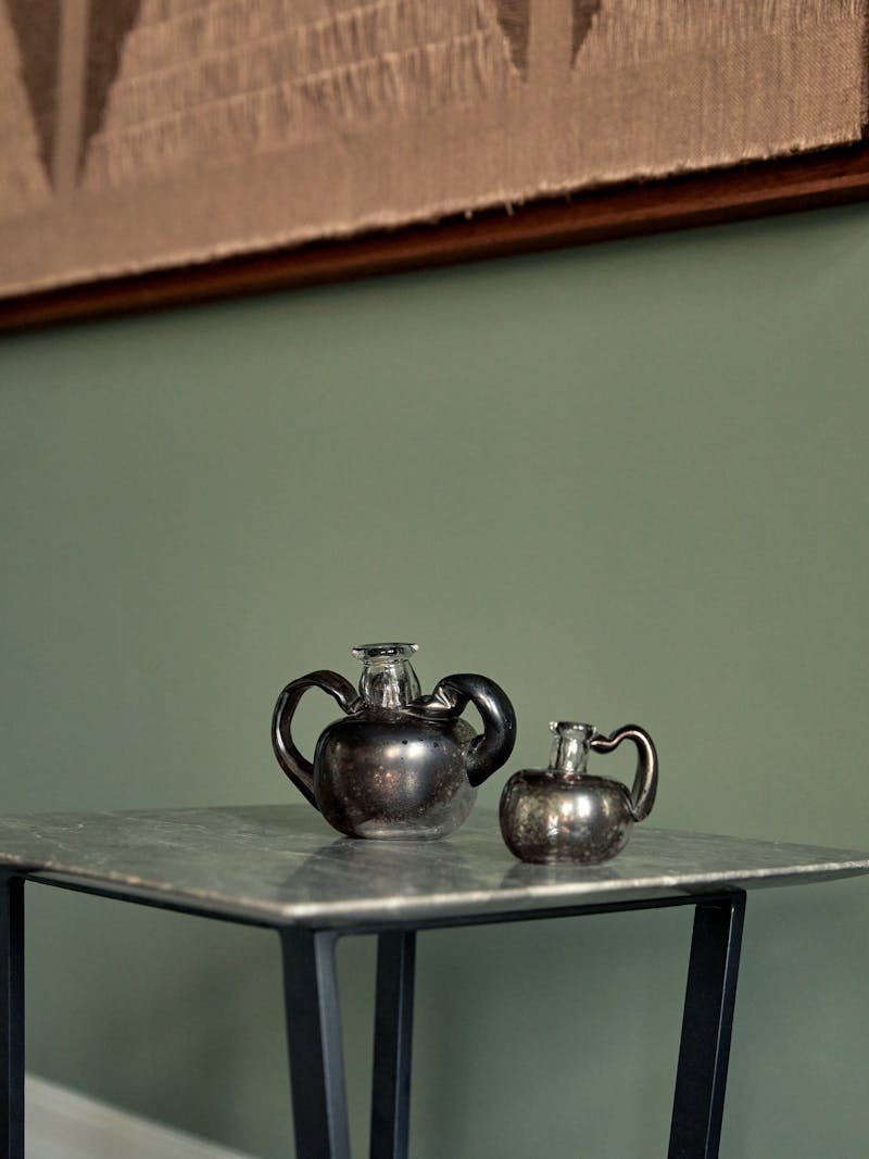 Two handblown glass vessels by French artist Pauline Bonnet sit on a marble table top in front of a green wall where the work of Sophie Rowley hangs. 