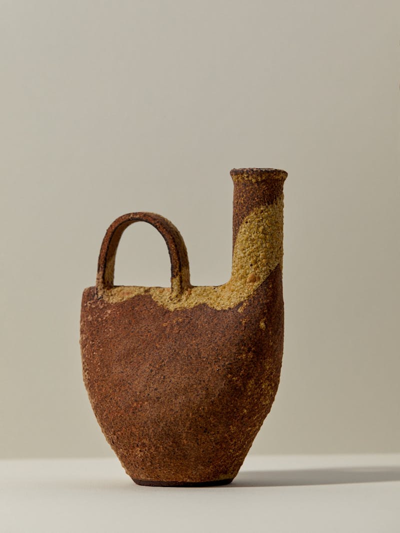 stoneware ceramic vessel by Catherine Dix with handle and pouring spout. the clay is fired in a combination of caramel, brown, cream and rust red colours. the sculptural vessel sits on a white table top.