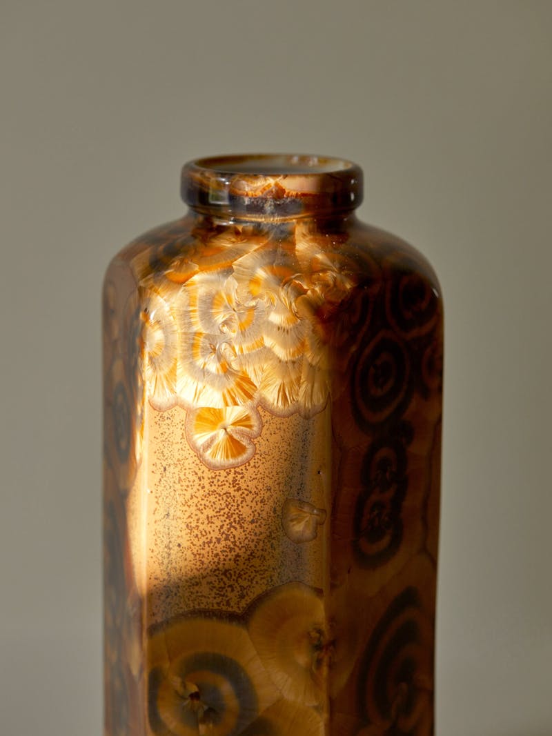 A tall hexagonal porcelain vase with a crystalline glaze in the colours of amber, gold, brown. The glaze has a natural burled pattern like a crystalline fossil or precious stone.