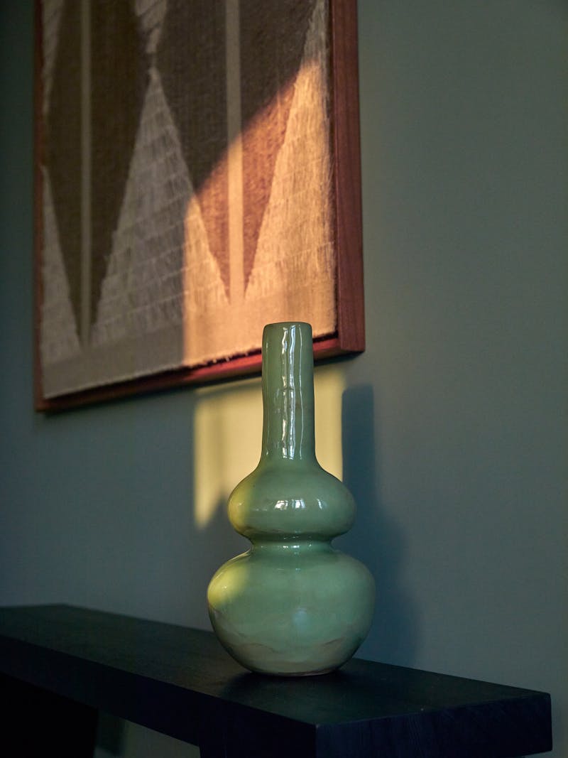 A curved green glazed ceramic stoneware vessel with a round base. The vase is made by Jade Patton, a South African ceramist and sits on a dark wooden bench. 