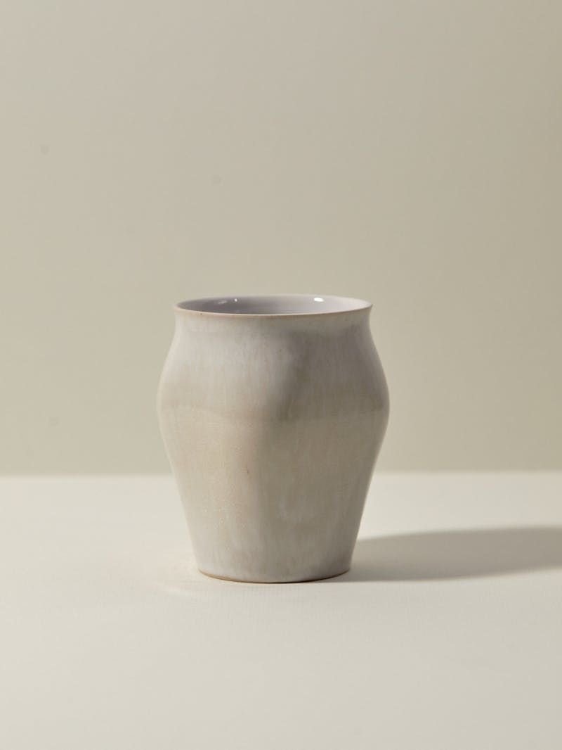 Unique handmade cream coloured ceramic curved cup from Russian designer Maria Tyakina. The vessel is slip cast and glazed and sits on a white table top.