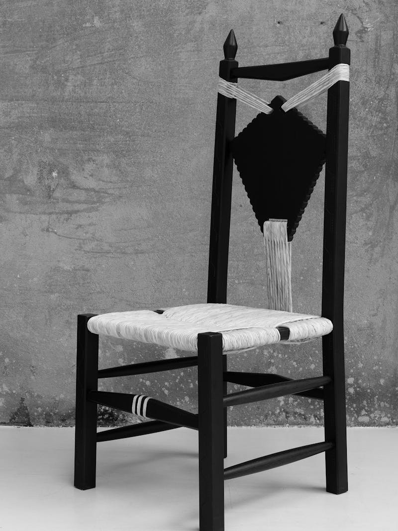 Black and white image displaying the Ines Chair from the 'scannu' series by Pretziada. The material is made from chestnut wood and contains woven beige-coloured decorative elements. This furniture object is part of the new 2024 spring collection 'Observations' by online gallery Metamorphoses Objects. 