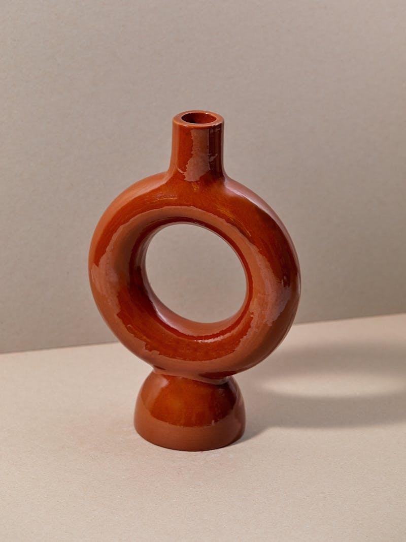 circular shaped round ceramic vase in terracotta with a clear glaze sitting on a white surface. The centre of the vase is open and the base of the vase is curved designed by Pretziada in Sardinia. 