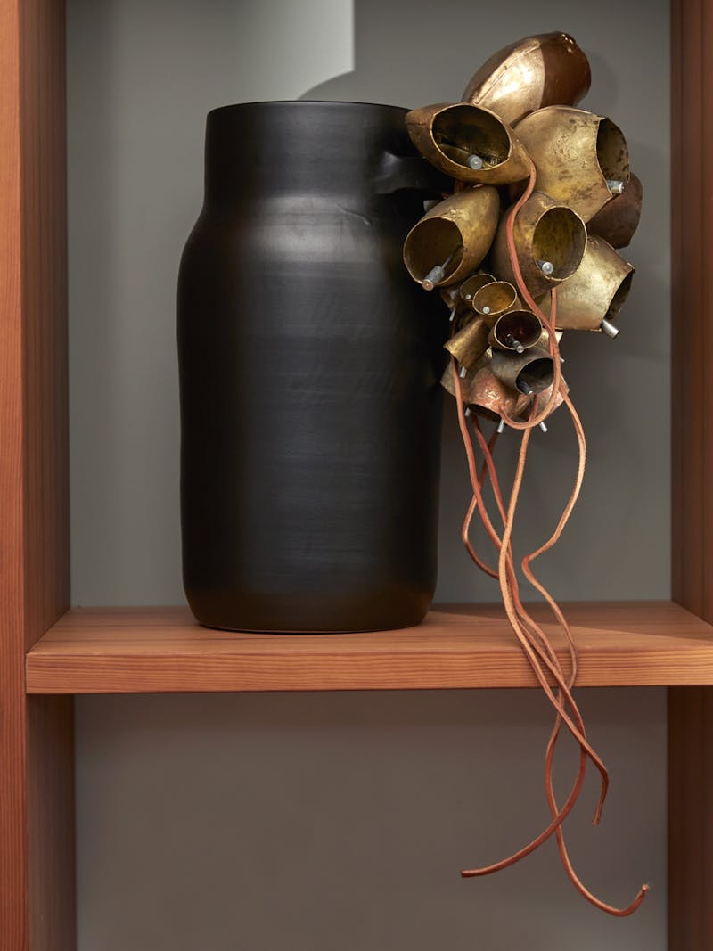 black matte terracotta vase with iron, leather and brass bells sitting on a wooden shelf vase made by Sardinian design duo Pretziada.
