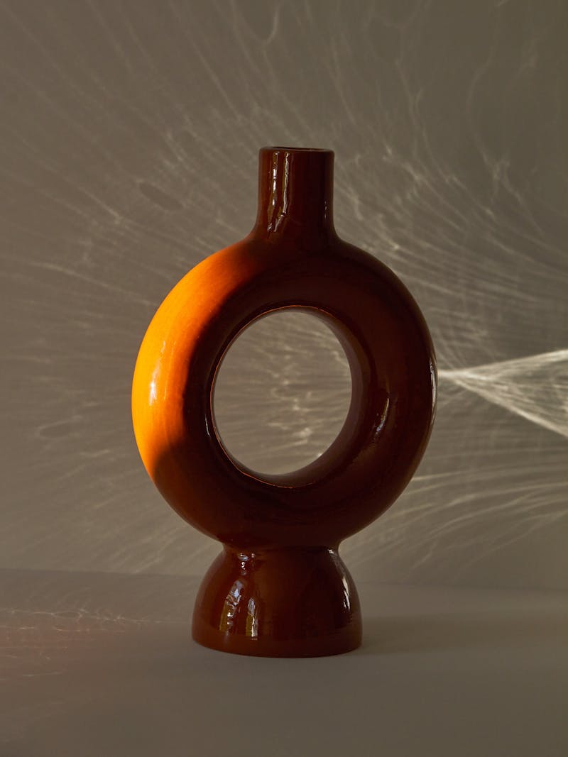 shadowy image of a circular shaped round ceramic vase in terracotta with a clear glaze. The centre of the vase is open and the base of the vase is curved designed by Pretziada in Sardinia. 
