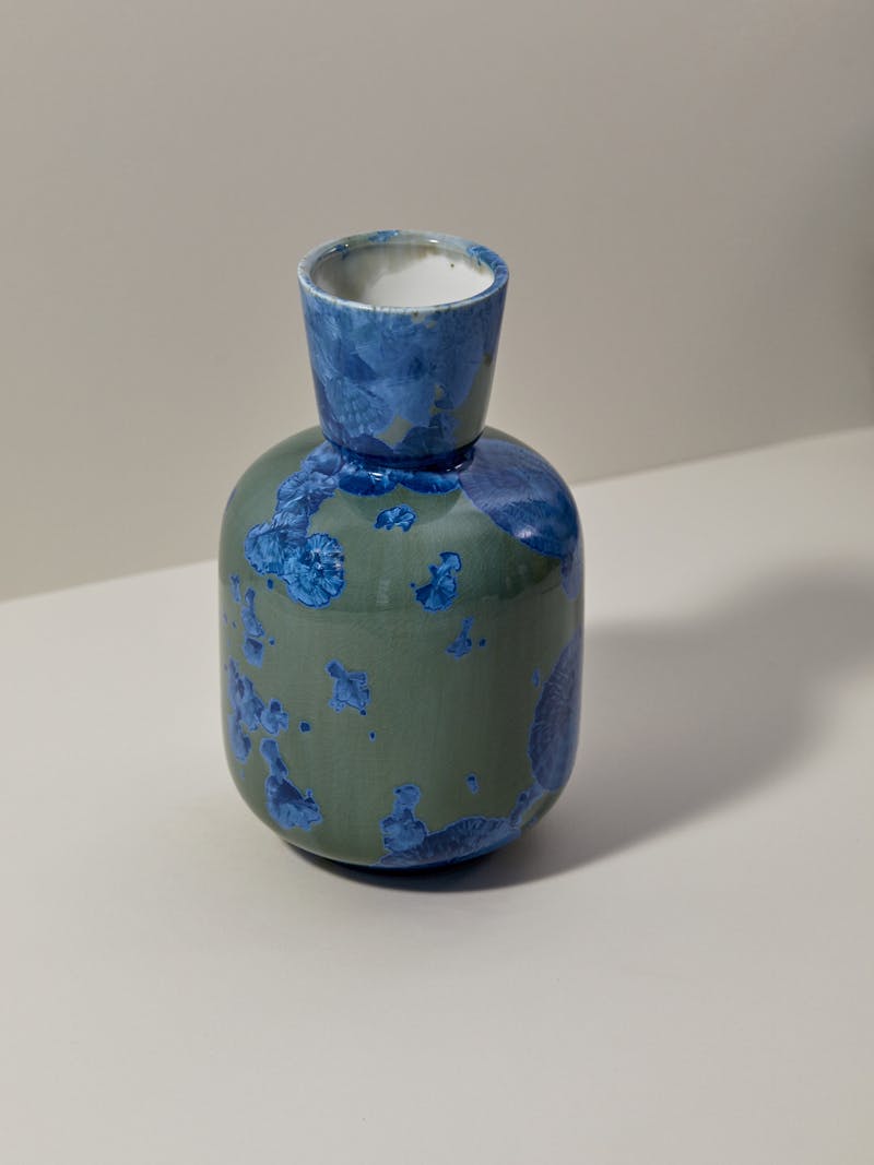 a sage green and sapphire blue porcelain vase with a round body and and tapered neck sits on a cream coloured table top. The vase has a crystal patterned glaze from natural materials. 